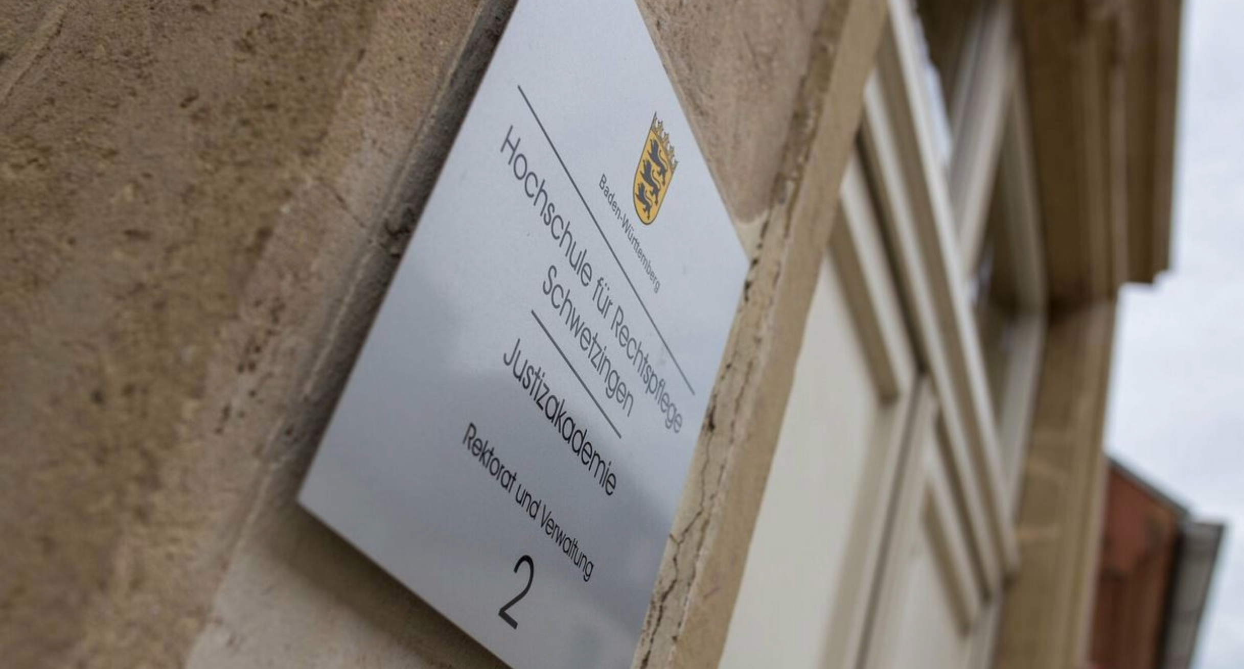 Silberfarbenes Schild an einem Gebäude. Das Schild trägt die Aufschrift Hochschule für Rechtspflege Schwetzingen Justizakademie Rektorat und Verwaltung.