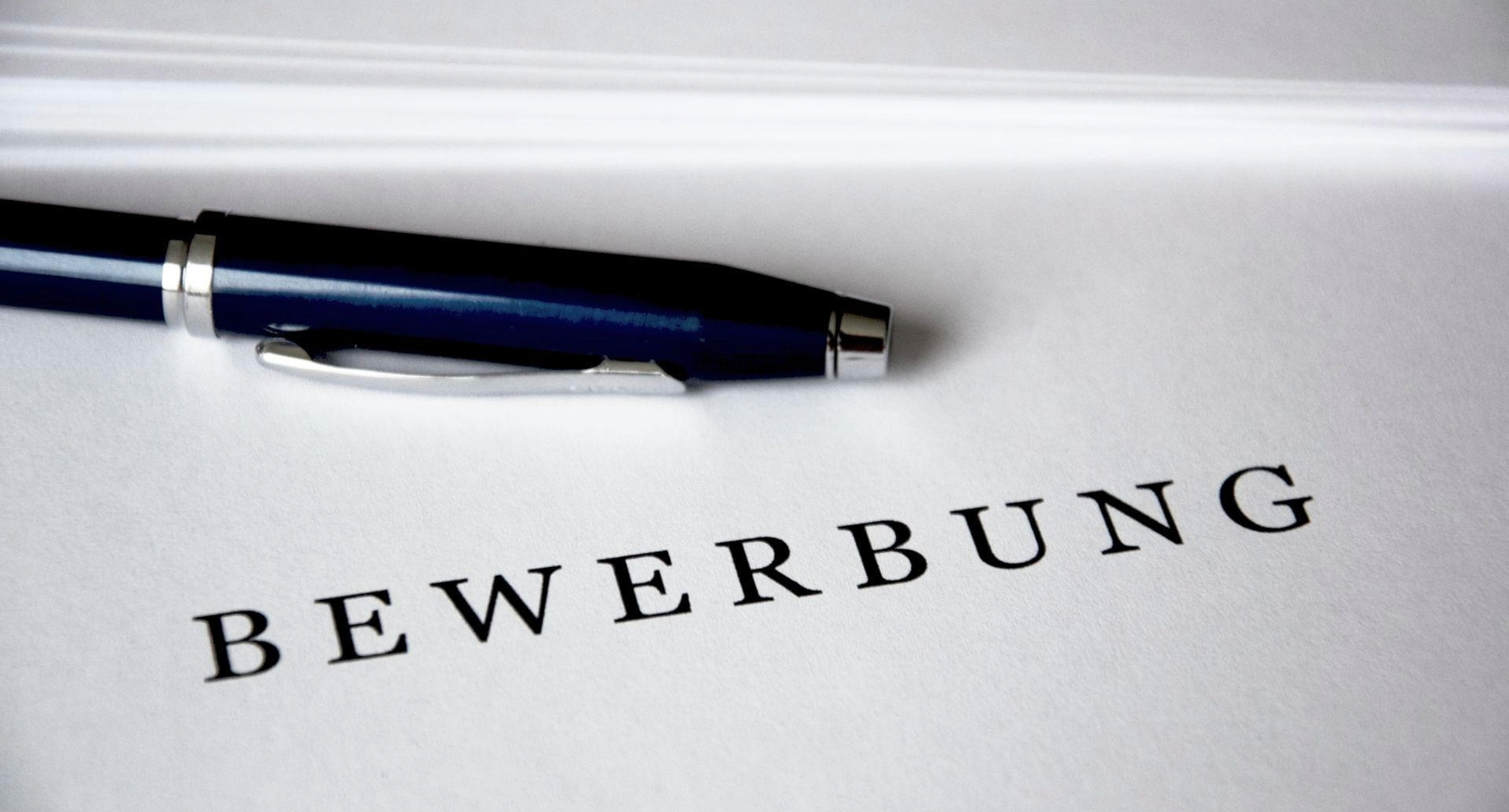 Das Bild zeigt einen Stift und ein Blatt Papier. Das Papier trägt den Aufdruck Bewerbung