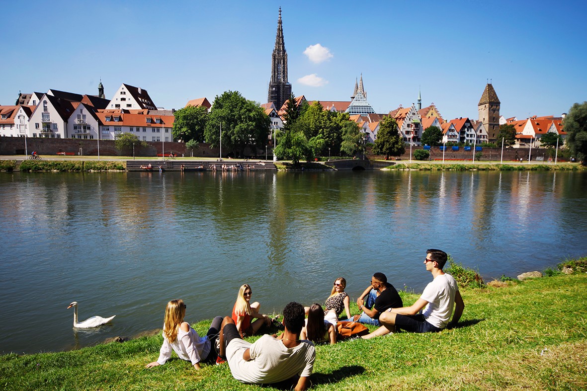 Bild zeigt junge Menschen die an der Donau sitzen. Im Hintergrund sieht man das Ulmer Münster 