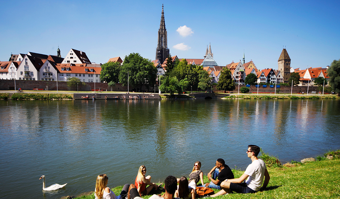 Junge Menschen die an der Donau sitzen. Im Hintergrund sieht man das Ulmer Münster 